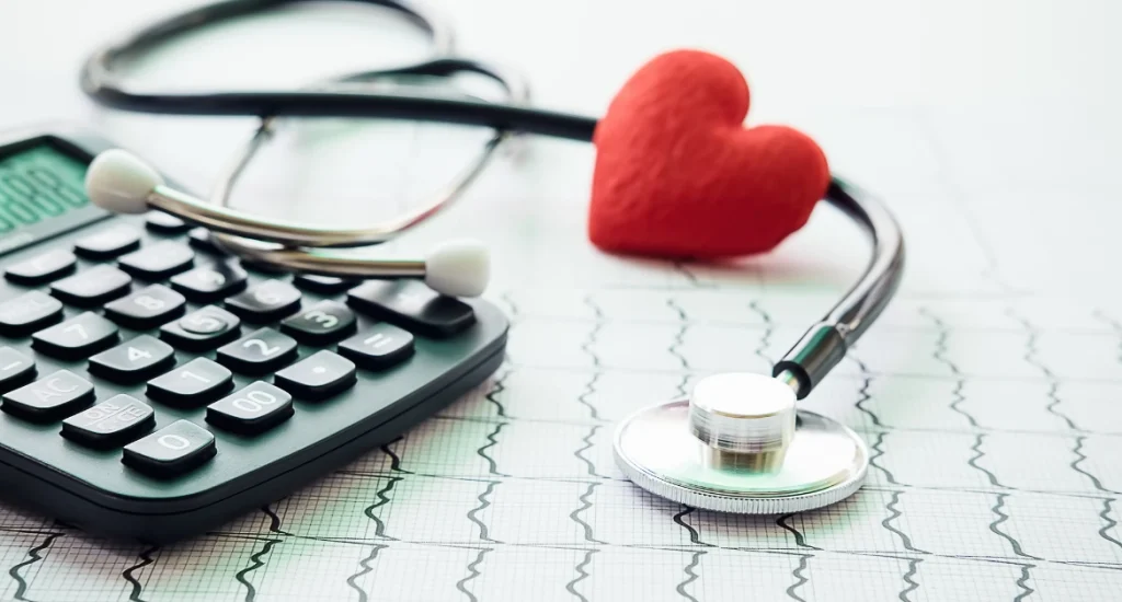 A stethoscope, calculator, and red heart model placed on an EKG chart, symbolizing the connection between medical care, heart health, and financial planning