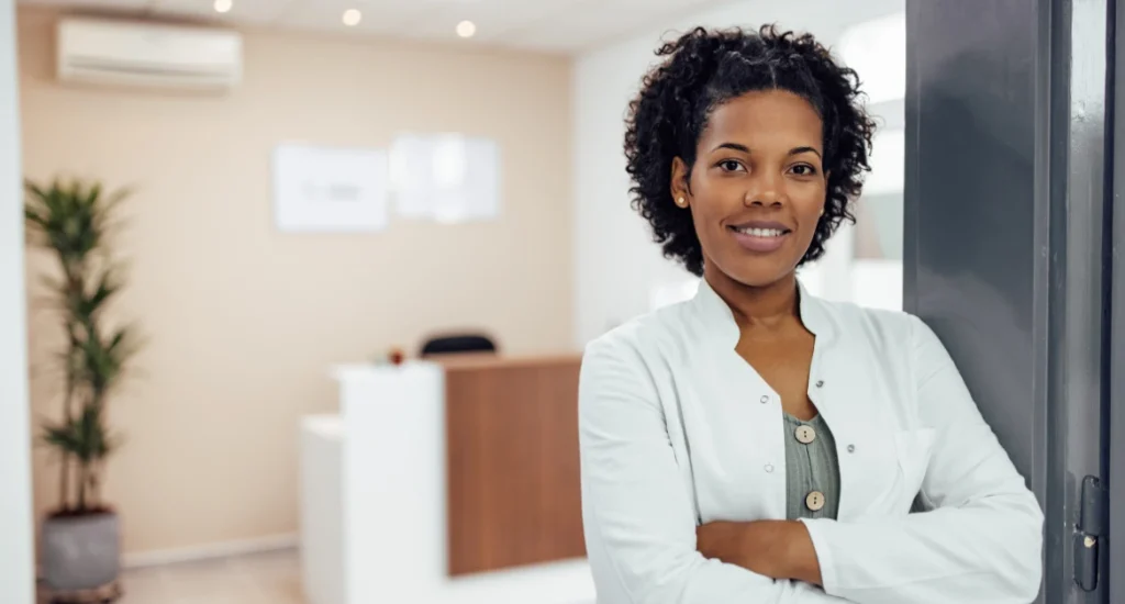 A smiling female healthcare professional in a white coat standing with arms crossed in a modern medical office or clinic reception area.